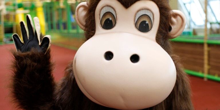 Monki-Maskottchen im Monki Park winkt vor einem Kletter- und Spielbereich des Indoor-Spielplatzes.